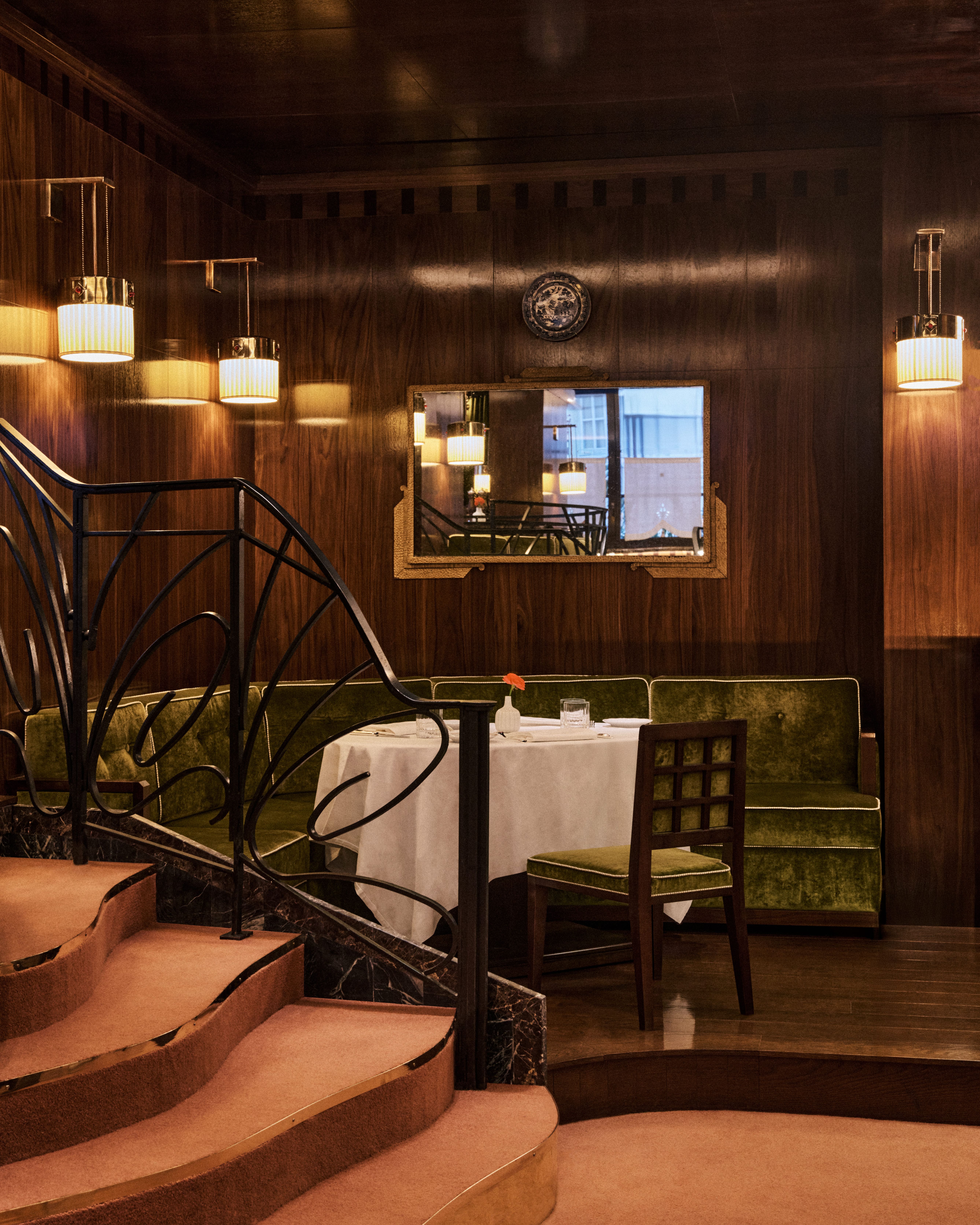 Intimate dining nook with green velvet banquette and white tablecloths, framed by curved pink-carpeted steps and black wrought-iron railing against warm wood-paneled walls.