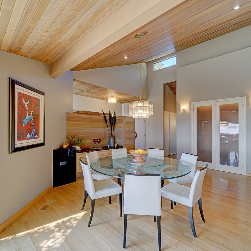 A well-lit dining room featuring a circular glass-top table with six white chairs. The room has a modern aesthetic with a sloped wooden-paneled ceiling, recessed lighting, and a striking contemporary chandelier. A framed piece of art hangs on the wall, and a wooden cabinet with a dark, reflective surface is visible to the left of the dining set. The floor is made of light-colored hardwood.