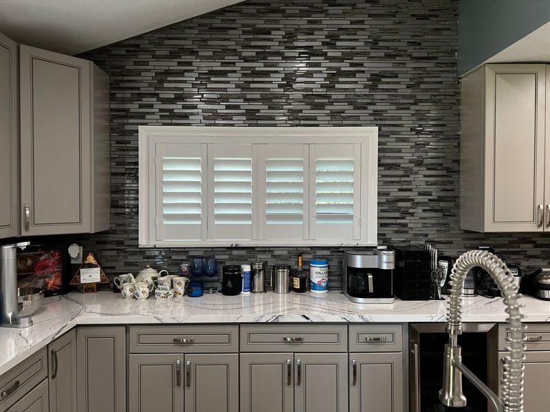 A modern kitchen featuring white plantation shutters that pop against a textured, dark gray tile backsplash. The space is finished with elegant gray cabinets, marbled white countertops, and professional-grade appliances.
