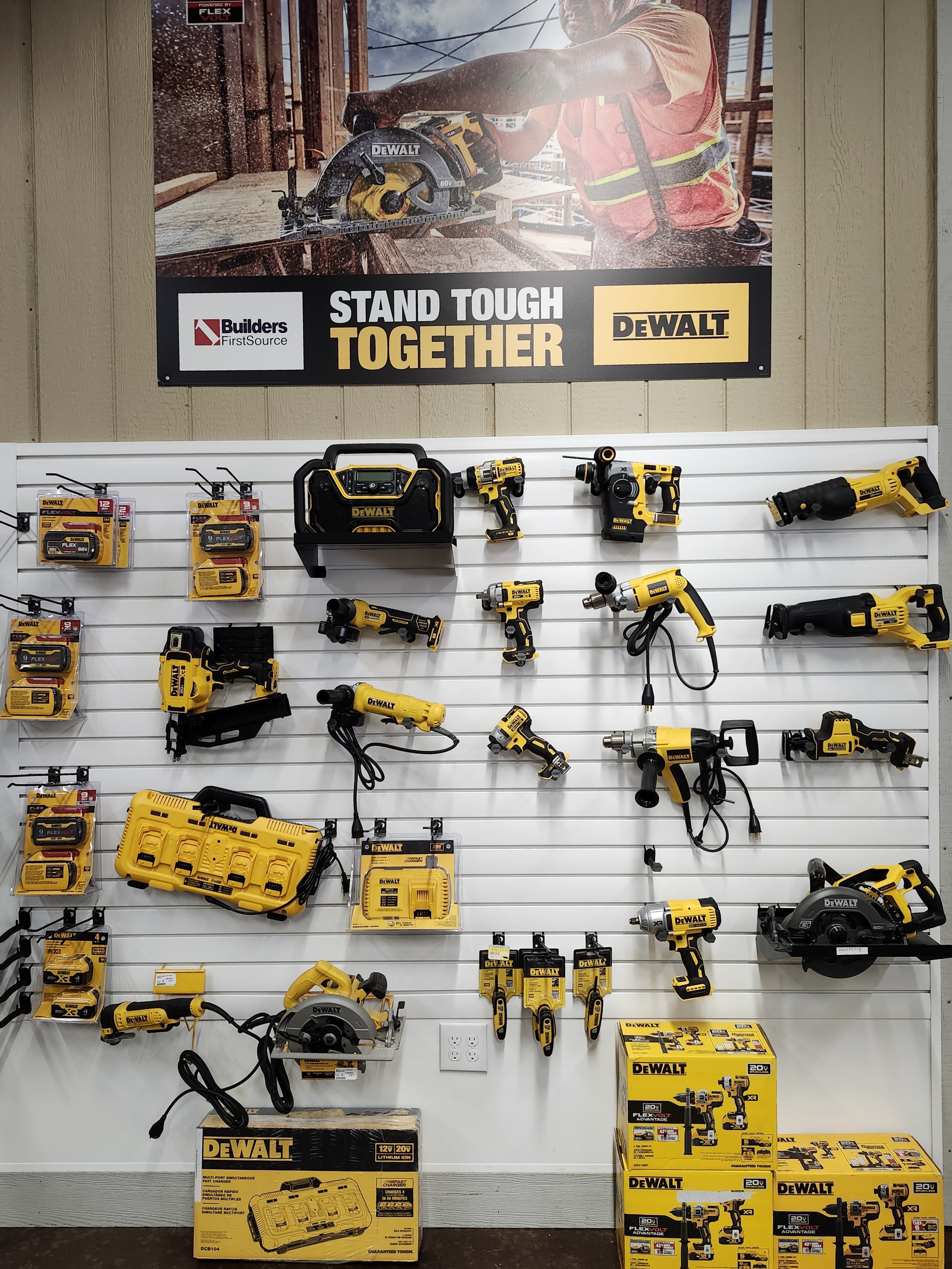 A comprehensive display of DeWalt power tools and accessories at a Builders FirstSource lumber yard. The display includes various drills, saws, nail guns, and battery packs, all neatly arranged on a slat wall. A prominent sign above the display reads "Stand Tough Together" showcasing the DeWalt brand.