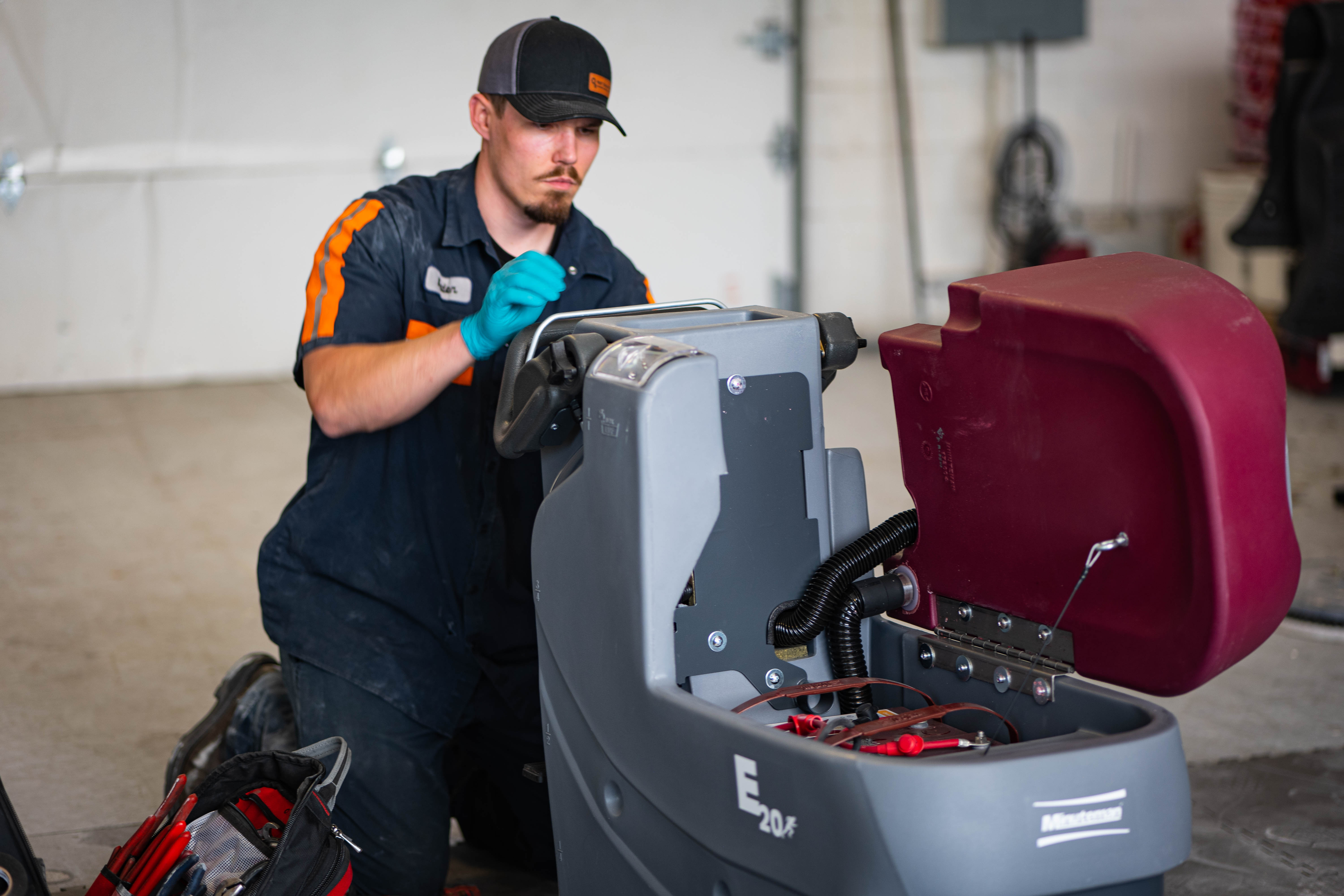 A WaySolutions technician kneeling in a service bay, performing a multi-point inspection on a Minuteman E20 walk-behind scrubber. The maroon recovery tank is propped open, revealing the battery bank and suction assembly. A professional tool bag is visible in the foreground, signifying a fully-equipped mobile or shop service call.