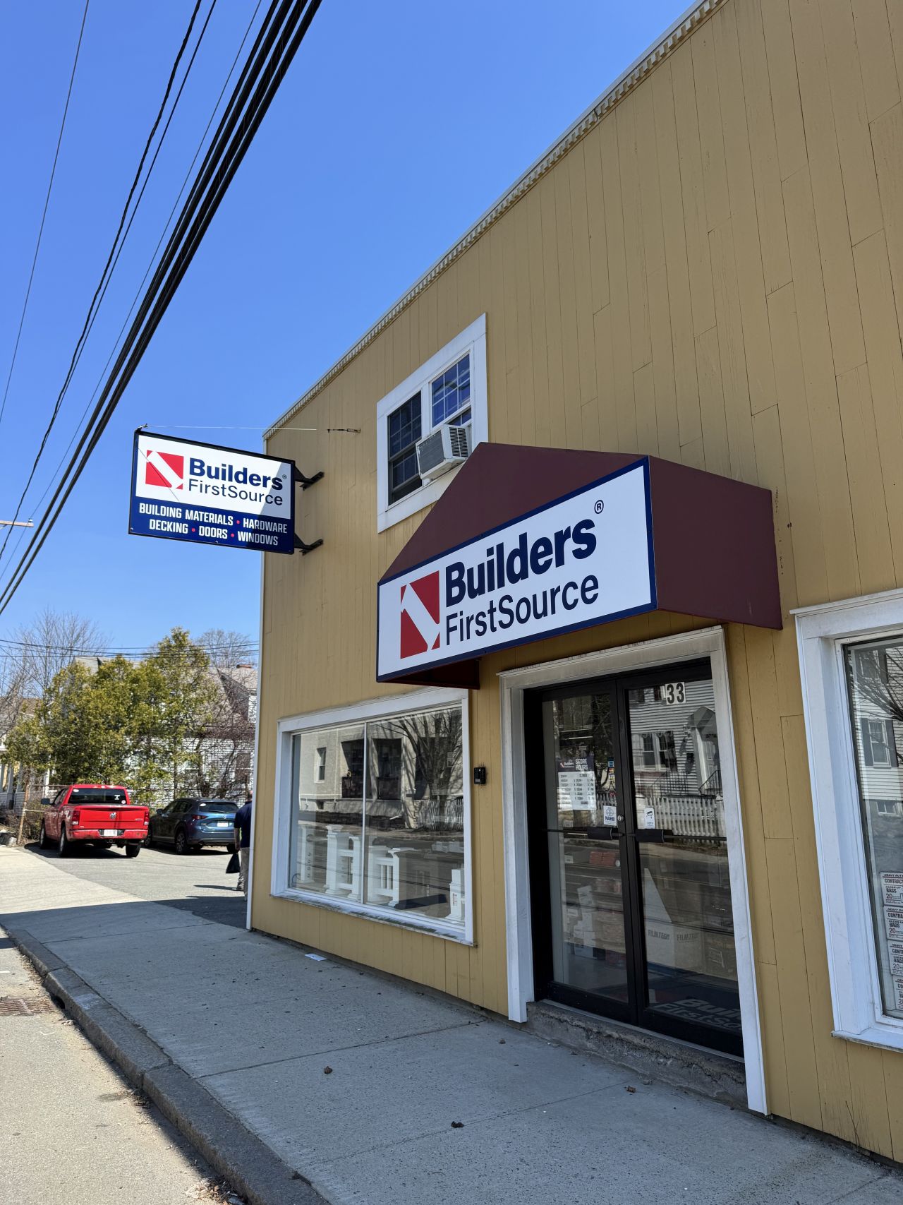Street‑level exterior of a Builders FirstSource building materials store featuring a yellow facade, front glass entrance, branded awning sign, and sidewalk access.