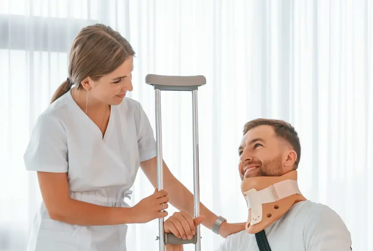 A medical professional in a white coat is assisting a man who is wearing a neck brace and a crutch. The man is smiling as the professional places her hand on his shoulder. The background is a brightly lit room with sheer curtains.