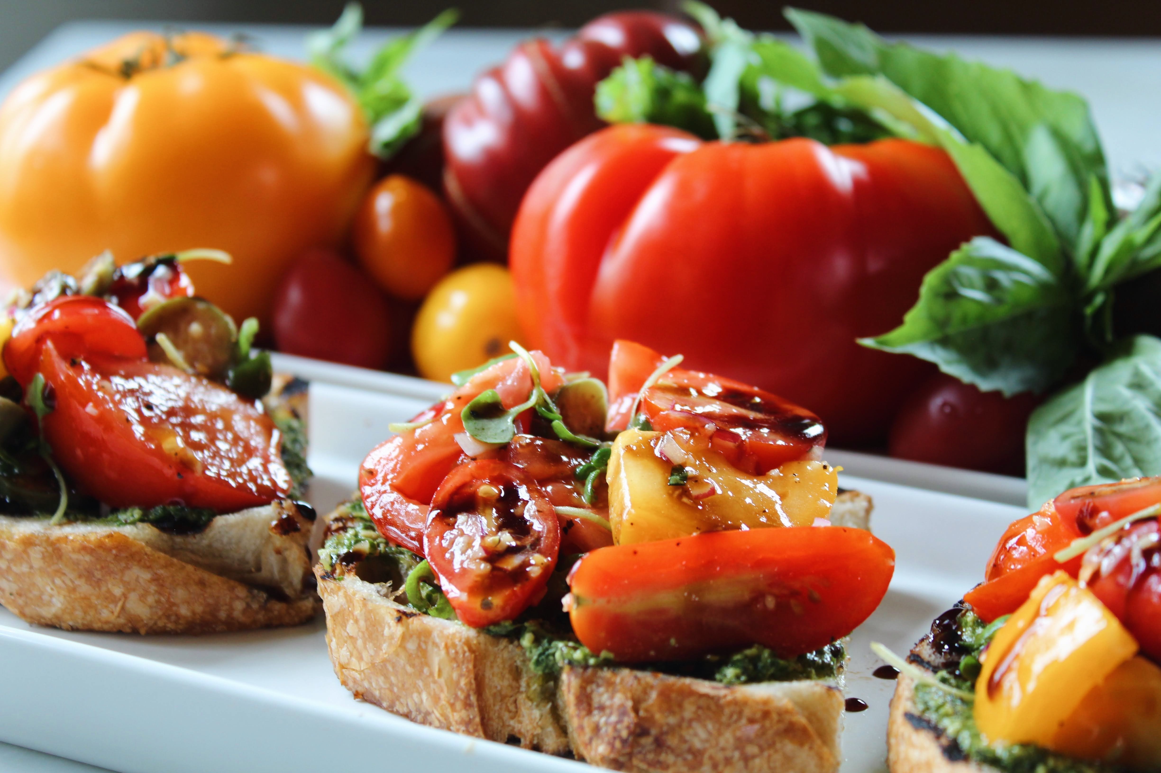 Tomato Bruschetta up close — marinated heirloom tomatoes and microgreens over basil pesto on grilled rustic sourdough, drizzled with balsamic glaze, with fresh heirlooms and basil in the background.