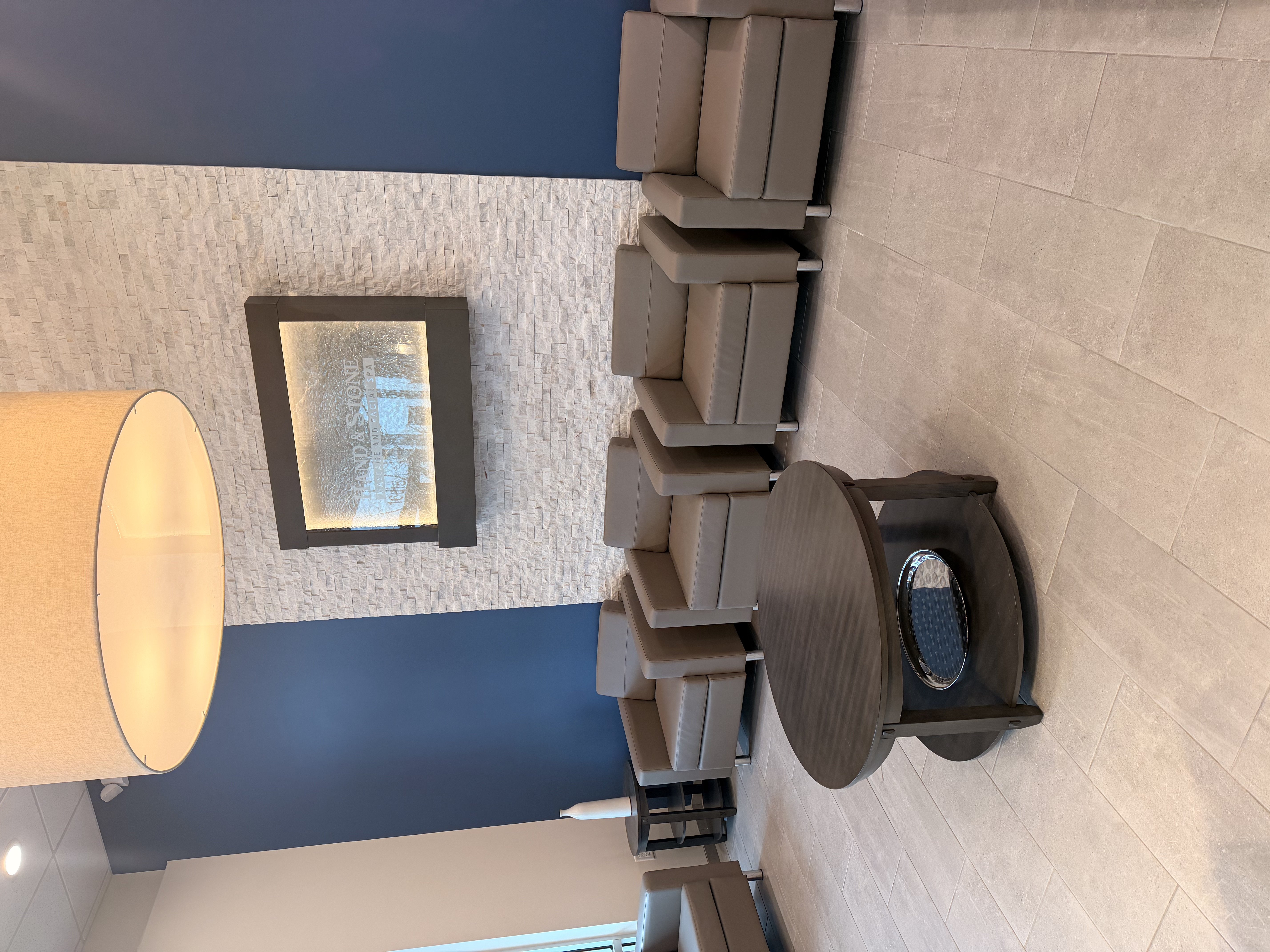 Waiting room with gray leather armchairs and round dark wood coffee table. Stacked stone feature wall with black-framed artwork. Navy blue accent wall with Hand & Stone logo partially visible. Recessed ceiling lights and gray tile flooring.