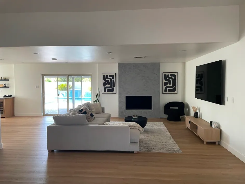 Spacious living room with modern design featuring large sectional sofa, wall-mounted television, minimalist fireplace, wood flooring, and sliding glass doors opening to a bright backyard, creating a clean, open-concept interior.