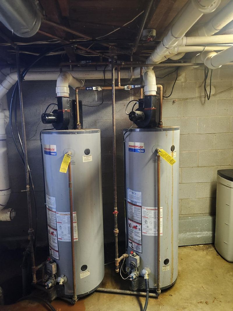 Two grey PowerFlex high-efficiency gas water heaters are installed side-by-side in a basement. The units feature top-mounted power vent blowers, copper water lines, and white PVC exhaust piping.