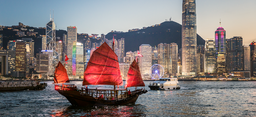 The Hong Kong skyline, lit up at night, with a traditional red-sailed junk boat in the foreground.