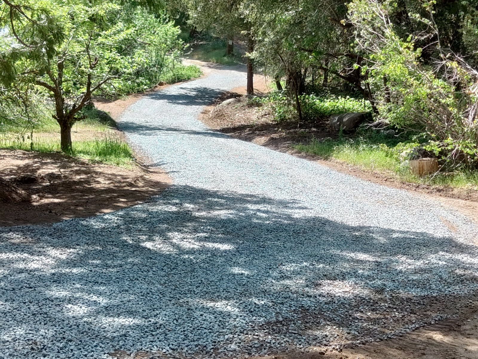 Curving gravel driveway extends through a shaded wooded property, covered with fresh crushed stone and bordered by trees, grass, rocks, and natural ground along the improved access route through landscape.