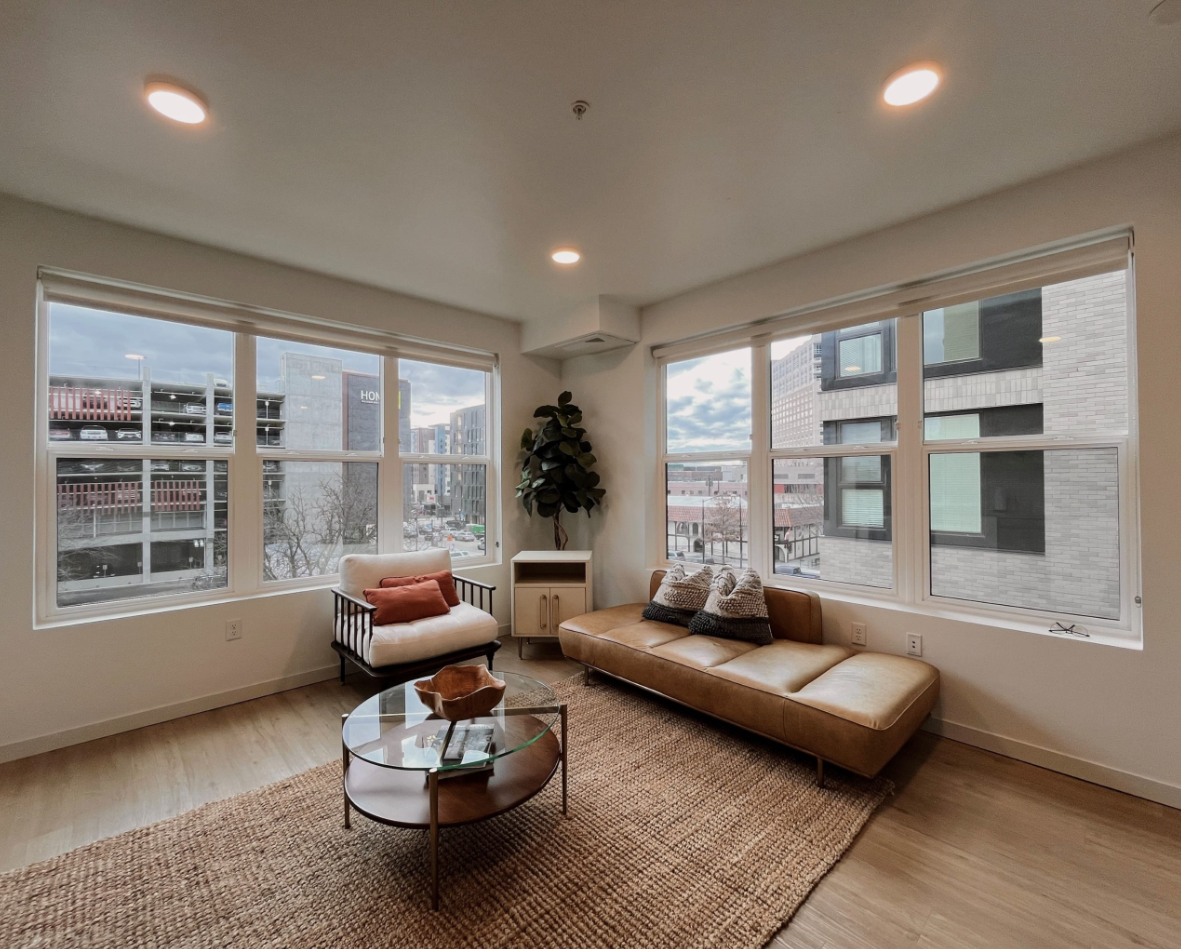 Spacious living area with a brown leather-style couch, jute-knotted rug, and two sets over oversized windows that overlook downtown Boise