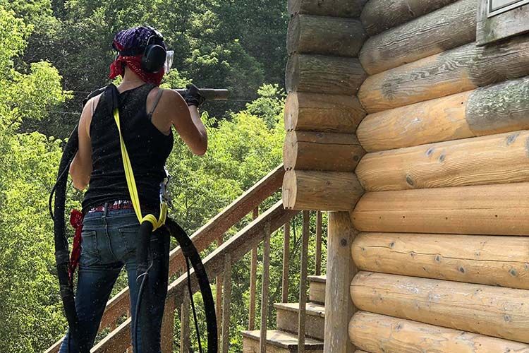 Technician sandblasting wooden cabin exterior, removing old finishes, dirt, and weather damage, restoring natural wood texture and preparing surface for staining, sealing, and long-term protection.