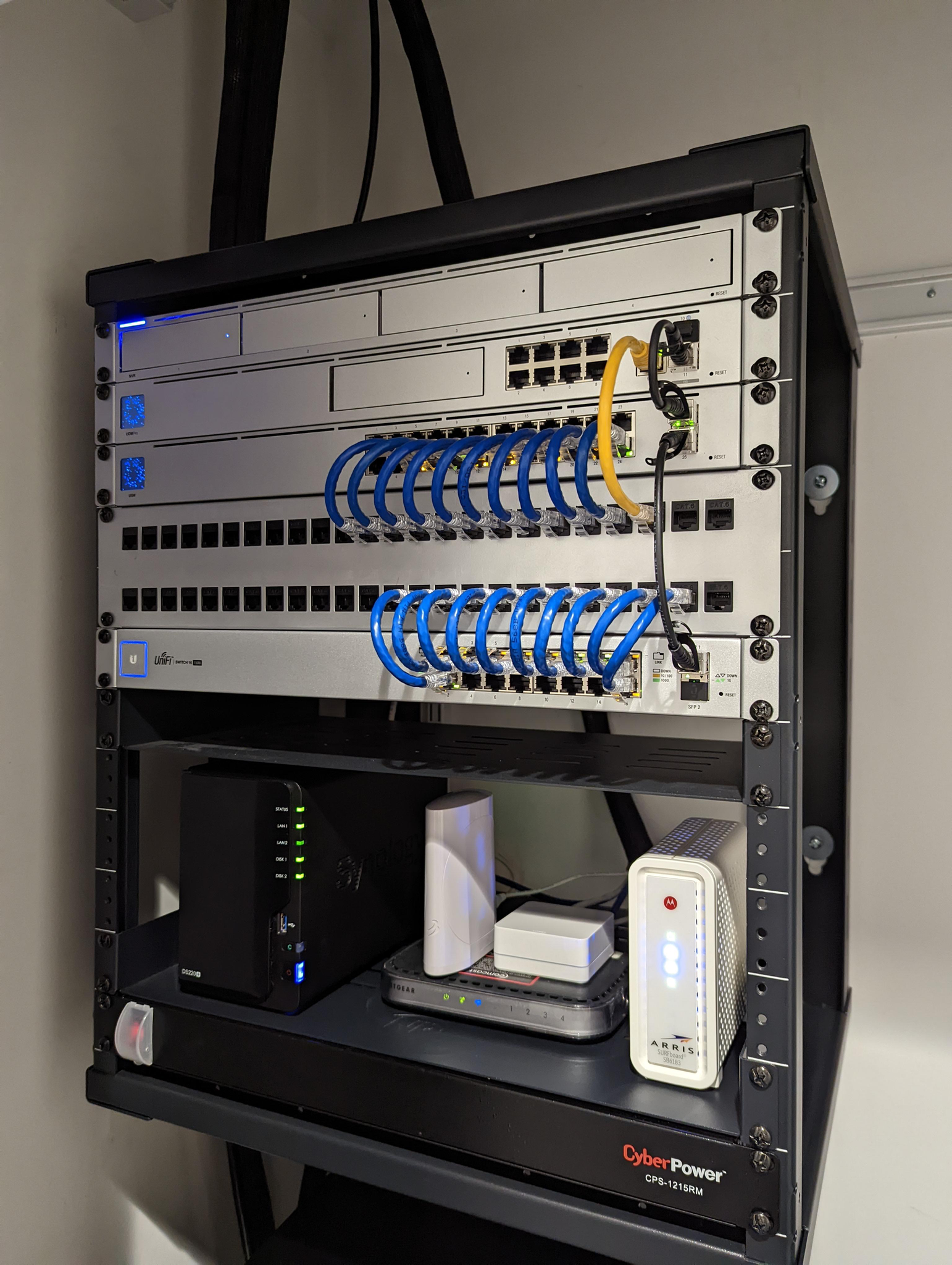 A network rack filled with various networking equipment including switches, routers, a NAS, and a modem. The equipment is organized and connected with blue and yellow Ethernet cables. The rack is mounted on a wall, and a power strip is visible at the bottom.