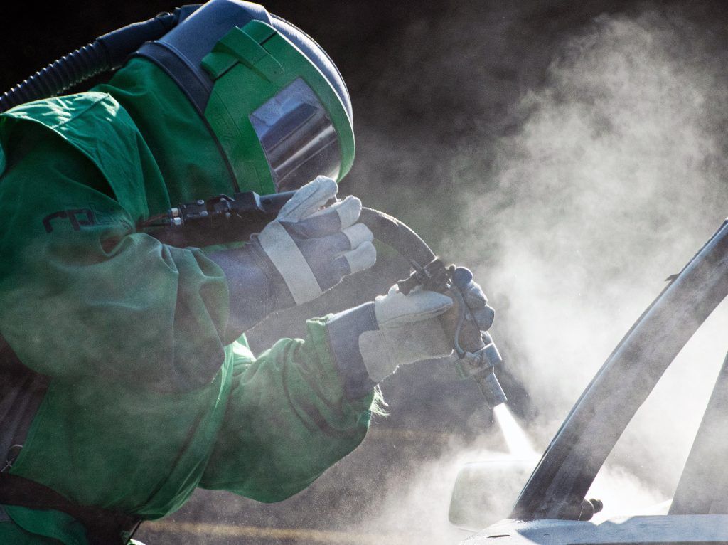 Close-up of technician performing abrasive blasting on metal surface, generating dust particles while removing coatings and contaminants, ensuring clean surface preparation for industrial finishing and protective coating applications.