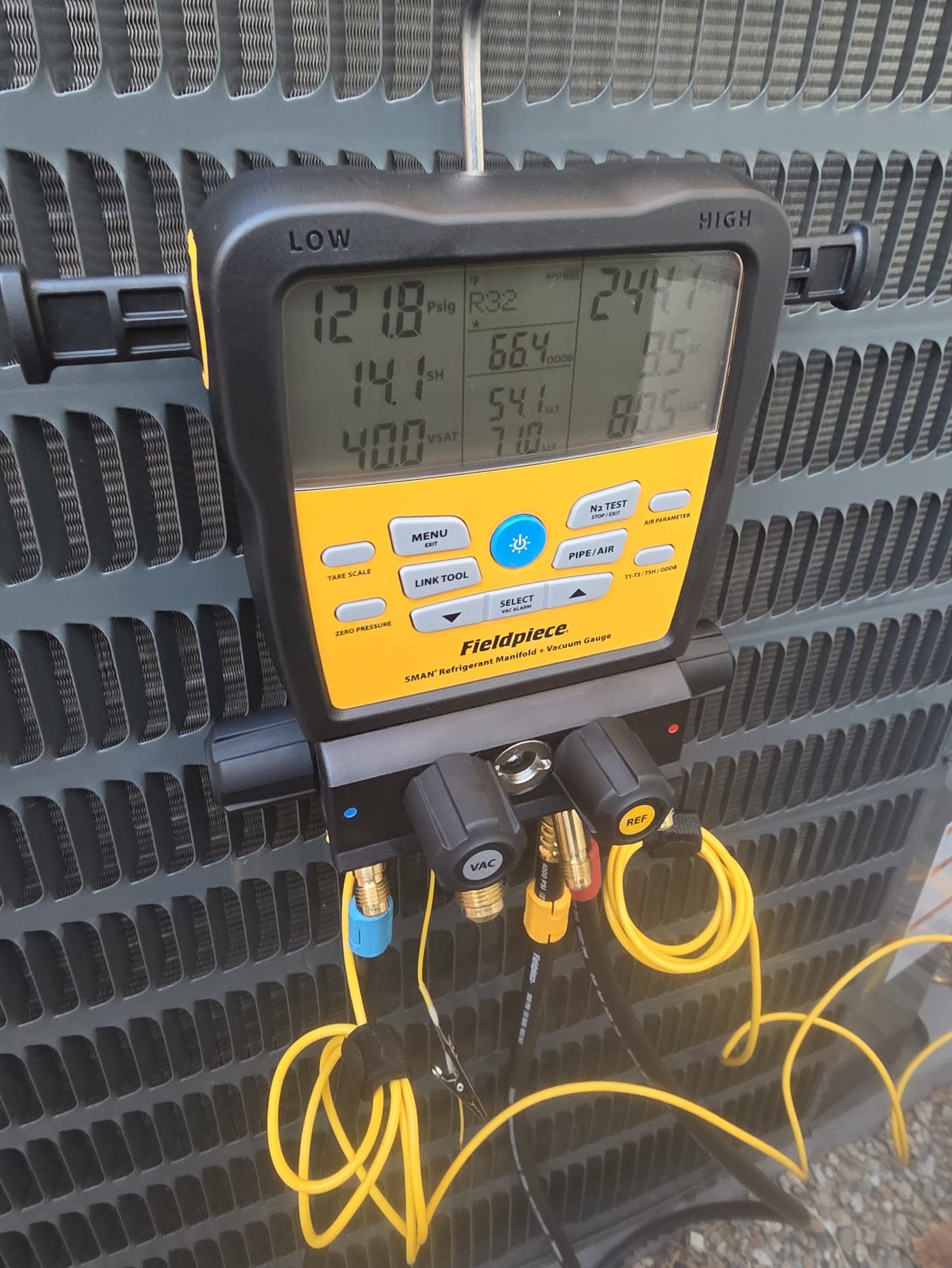 A Fieldpiece SMAN refrigerant manifold and vacuum gauge attached to an AC condenser, displaying live refrigerant pressure and system performance readings during a service call.