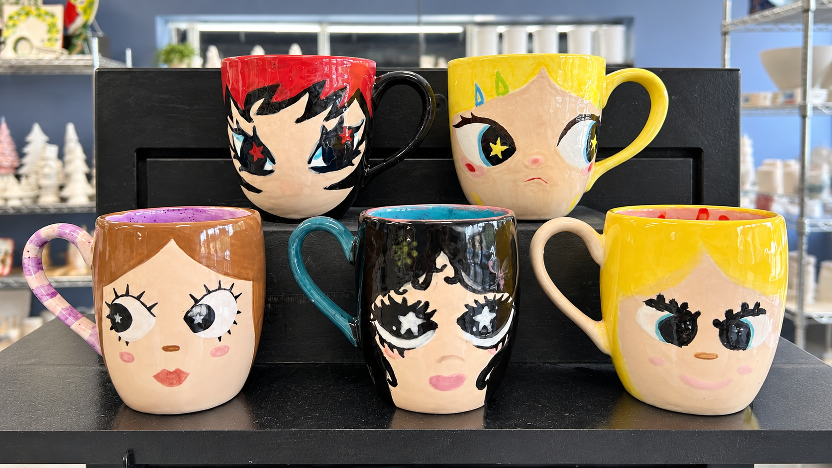 Colorful hand-painted face mugs displayed at Do It Yourself Crafts. Visit the pottery studio to design a unique mug that adds creativity and cheerful character to every coffee or tea break every day!