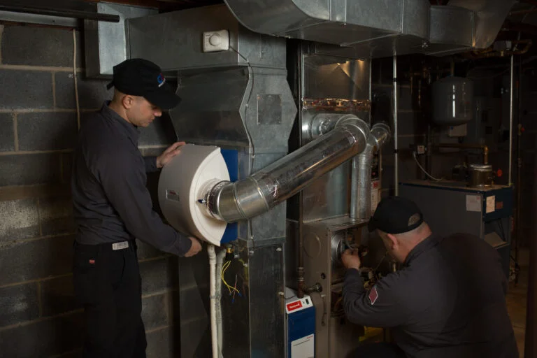 Two men in work uniforms are in a basement, working on a furnace and a humidifier. One man is on the left, holding a white humidifier unit and attaching it to the ductwork. The other man is on the right, kneeling down and working on the furnace with tools. The basement has a concrete block wall and visible pipes and ductwork.