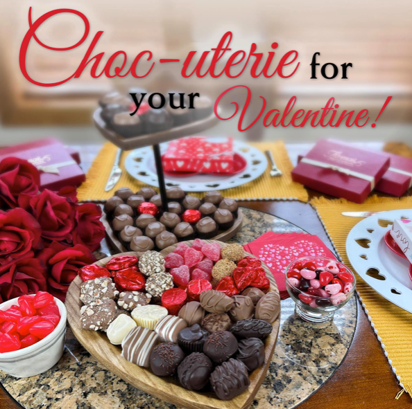 This photograph captures a creative and appealing Valentine's Day chocolate arrangement from Florence's exquisite chocolates, likely taken in their Rexburg, Idaho location. The image promotes a "Choc-uterie" board, a play on "charcuterie," designed for sharing with a loved one. The heart-shaped board is packed with a diverse selection of chocolates and candies, showcasing the variety of treats offered. The arrangement includes recognizable items like chocolate-covered pretzels, truffles with different toppings, neatly wrapped chocolate bars, and an assortment of colorful candies in red, pink, brown, and white. The surrounding elements, such as the red roses, heart-shaped chocolate boxes, and the small bowl of red and white candies, further enhance the Valentine's Day theme and create a visually appealing composition.