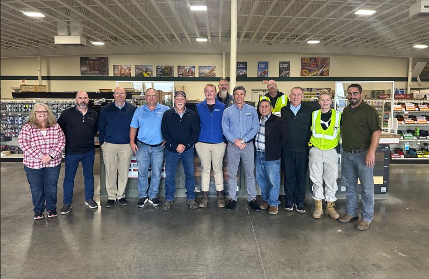 A group of 15 Builders FirstSource employees and partners stands together inside a well-lit retail or warehouse space. They’re gathered in front of store shelving stocked with hardware and building supplies, smiling for a group photo beneath a high industrial ceiling.
