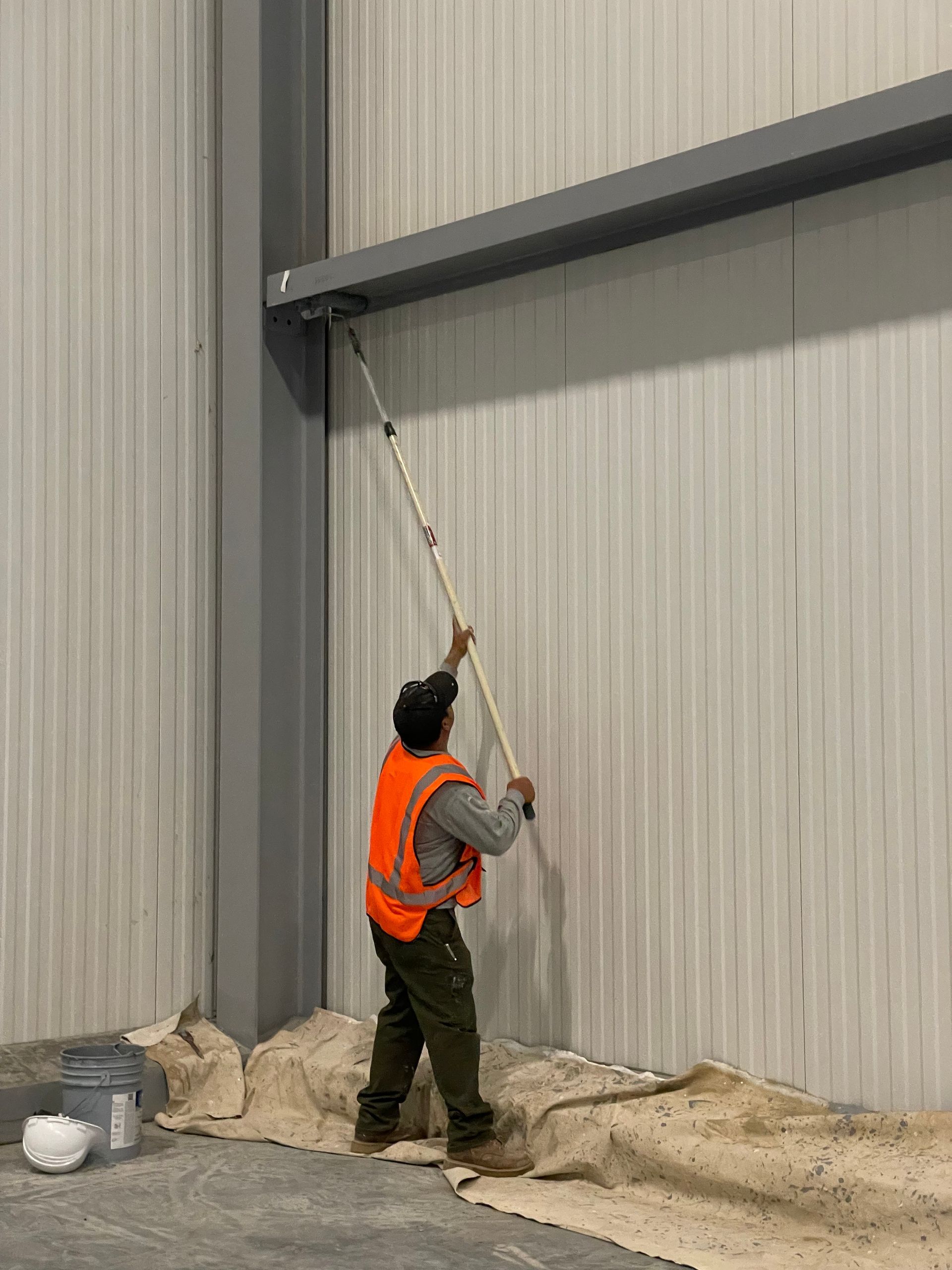 A worker wearing a safety vest paints a large industrial wall using a roller on an extension pole, with protective coverings on the floor, showing active maintenance inside a warehouse space.