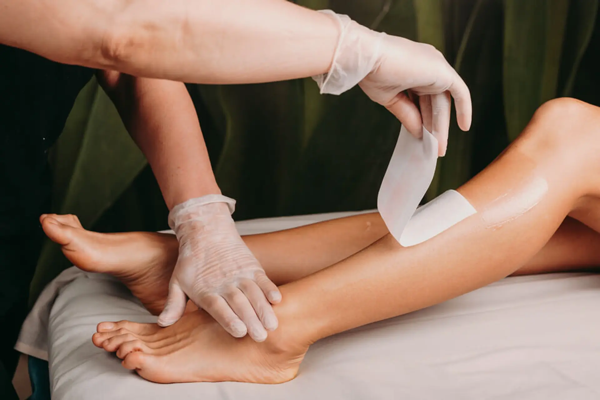 A close-up shot shows a person's legs and feet as a professional, wearing white gloves, applies a waxing strip to one of the legs. The setting appears to be a clean and professional spa or salon, with a white sheet covering a surface and a dark green fabric in the background.
