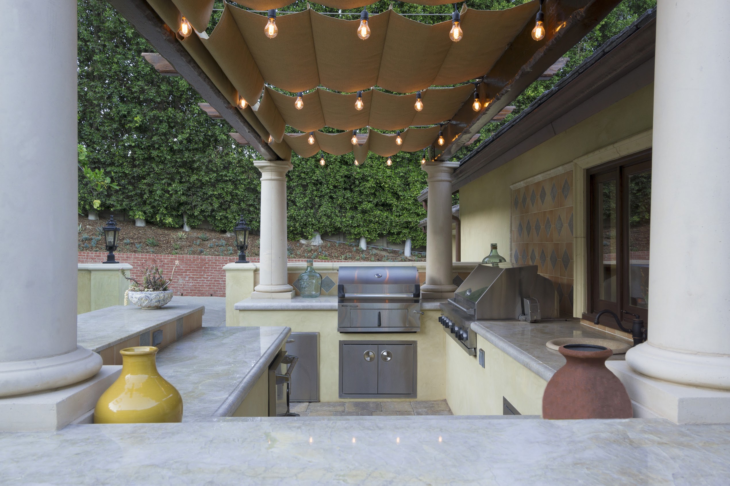 Elegant outdoor kitchen with marble counters, grill, urns, columns, and string lights beneath a canopy, creating a warm, inviting patio.