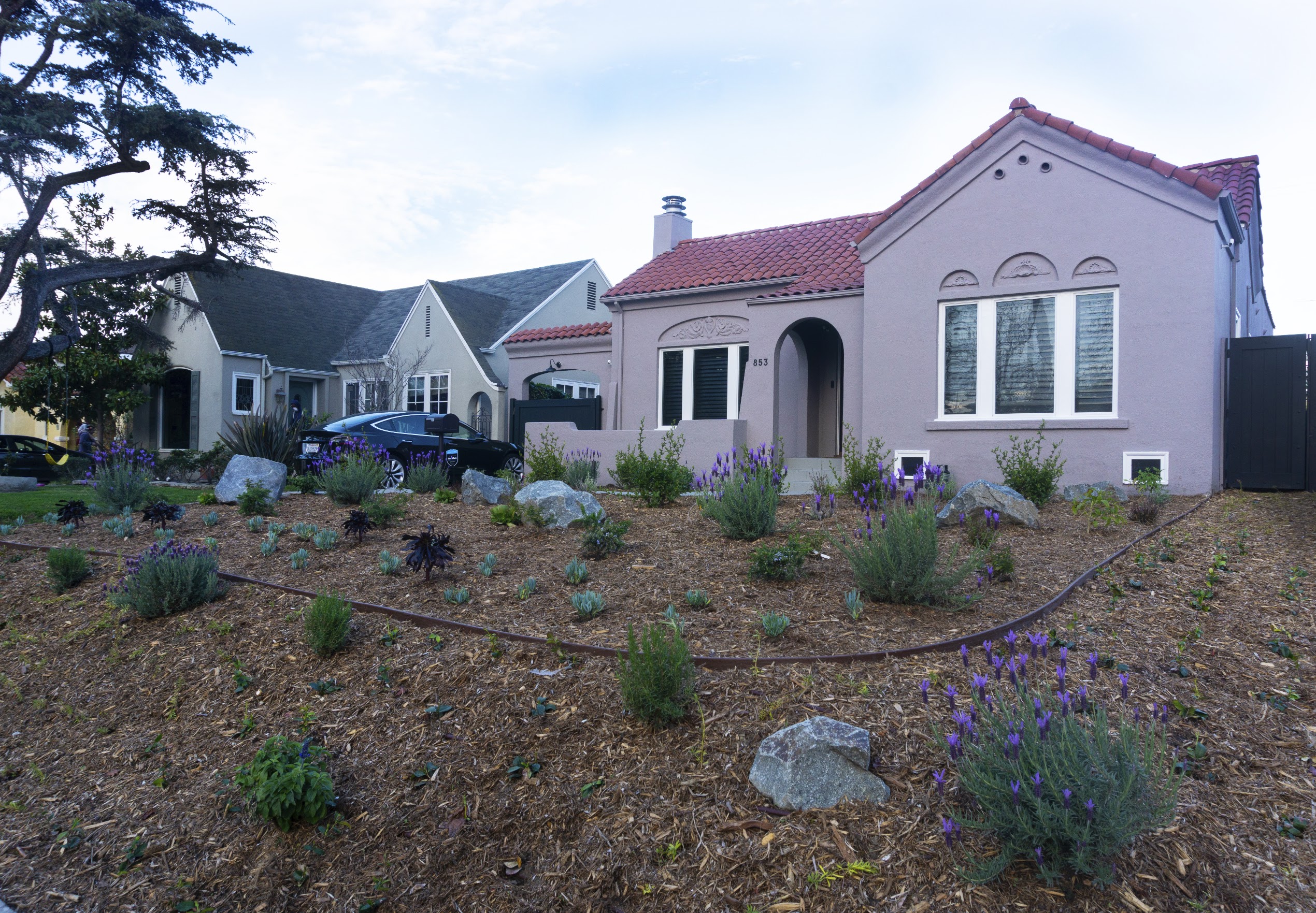 Charming pink stucco home with red tile roof and arched entry, surrounded by lavender and drought-tolerant landscaping under blue skies.