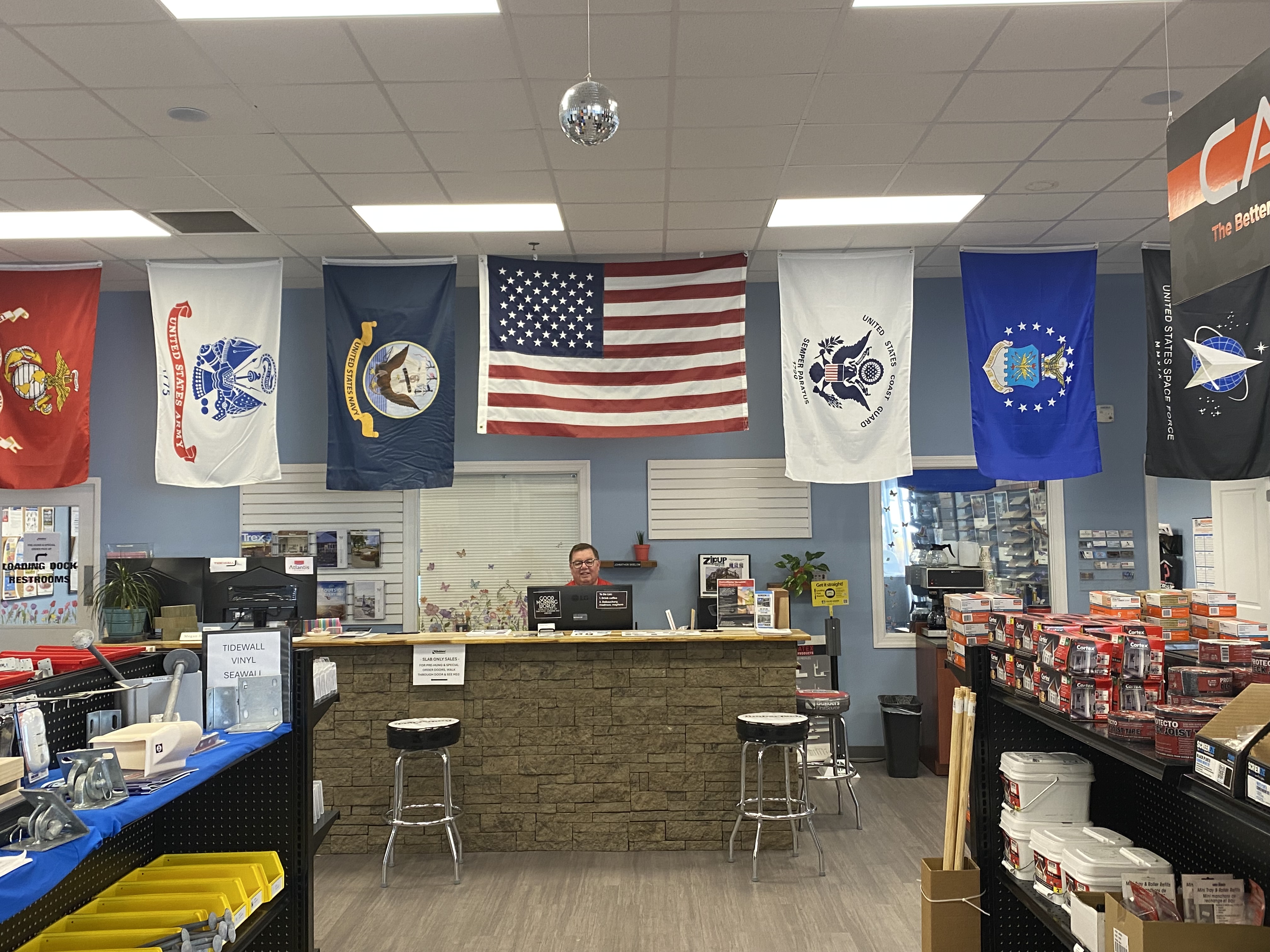 The front counter at Builders FirstSource Chesapeake location, adorned with multiple American and military service flags. The customer service desk is stocked with essential building materials, fasteners, and hardware supplies, reflecting a commitment to quality construction and support for service members.