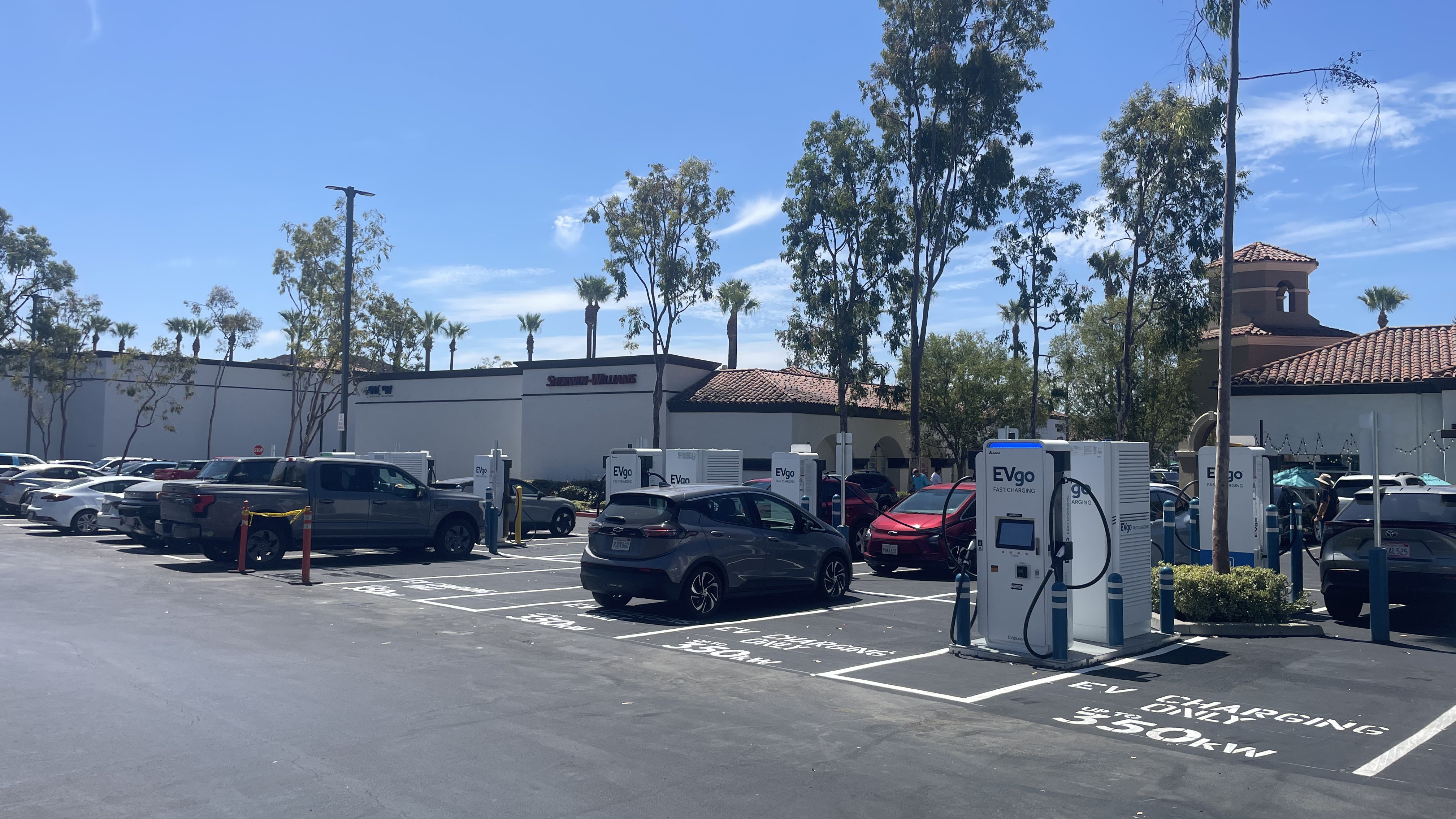 Several EVgo fast charging units installed in marked parking spaces at a public charging station, with EVgo logos on the chargers, protective bollards around each unit. NACS and CCS chargers featured in the image.