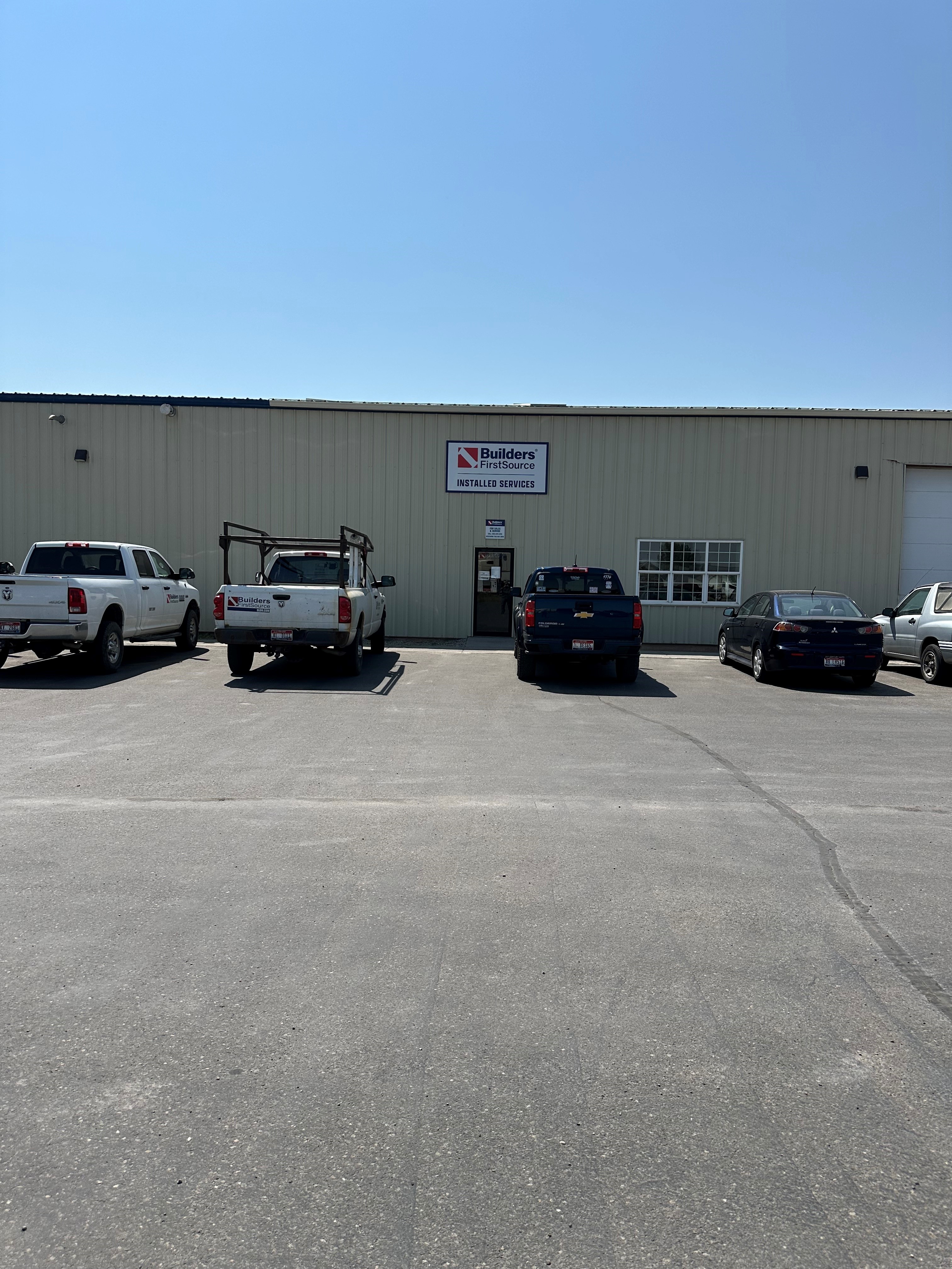 A Builders FirstSource facility in Idaho Falls, specialized in window and garage door installation services, with a parking lot in front featuring a white company truck and two other vehicles.