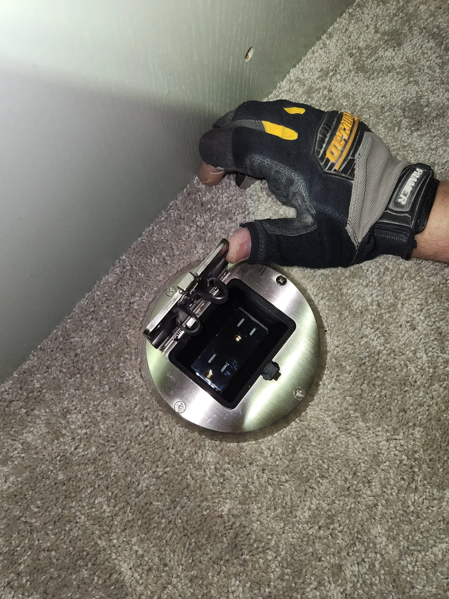 A gloved hand is shown reaching towards a newly installed floor outlet with a brushed metal finish. The outlet is recessed into a beige carpeted floor next to a light green wall. The outlet cover is open, revealing two black electrical sockets. A screw is visible on the wall just above the outlet.