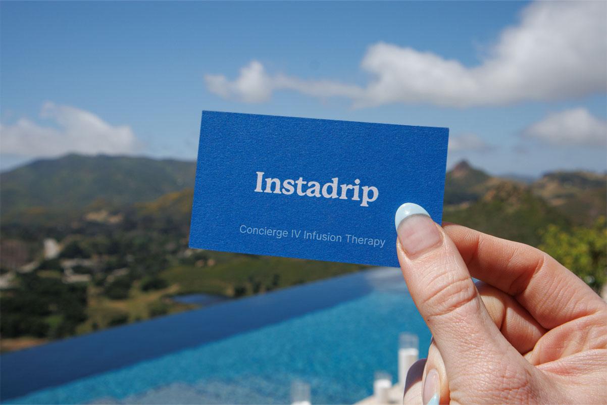 A close-up of a hand with light blue nails holding a blue business card that reads "Instadrip – Concierge IV Infusion Therapy," set against a scenic mountain backdrop with a clear sky and an infinity pool below.