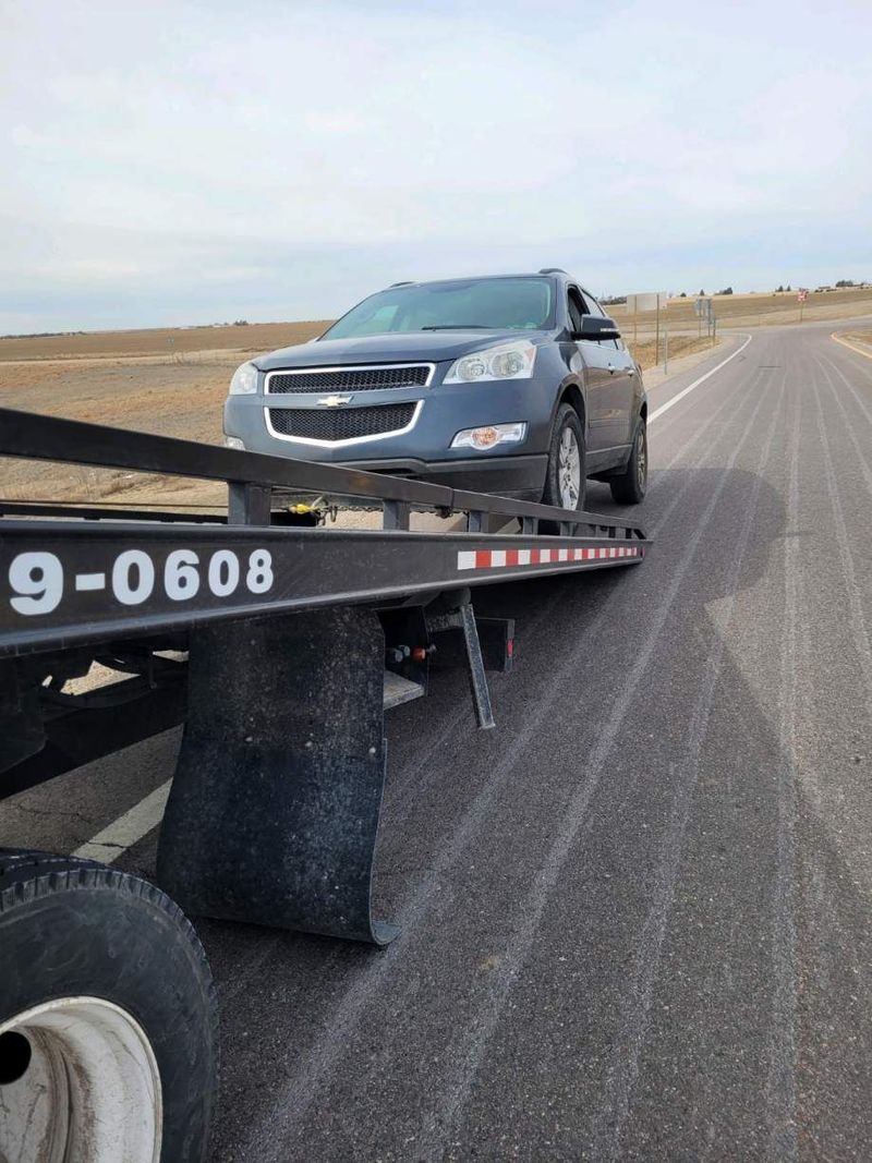 A close low angle photo shows a dark gray Chevrolet SUV sitting atop the angled ramp of a flatbed tow truck, parked beside an asphalt road in a rural landscape.