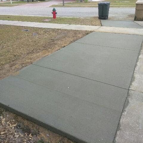 Freshly poured residential concrete sidewalk section connecting driveway to street, smooth finish with control joints, bordered by soil and grass, near red fire hydrant and curbside trash bin.