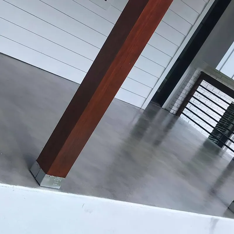 A close-up, angled shot of a modern architectural space. A thick, dark wood beam extends diagonally across the frame, resting on a polished concrete floor. The background features a white wall with horizontal plank detailing and a section of brickwork with a black metal railing or grate. The overall aesthetic is minimalist and contemporary.