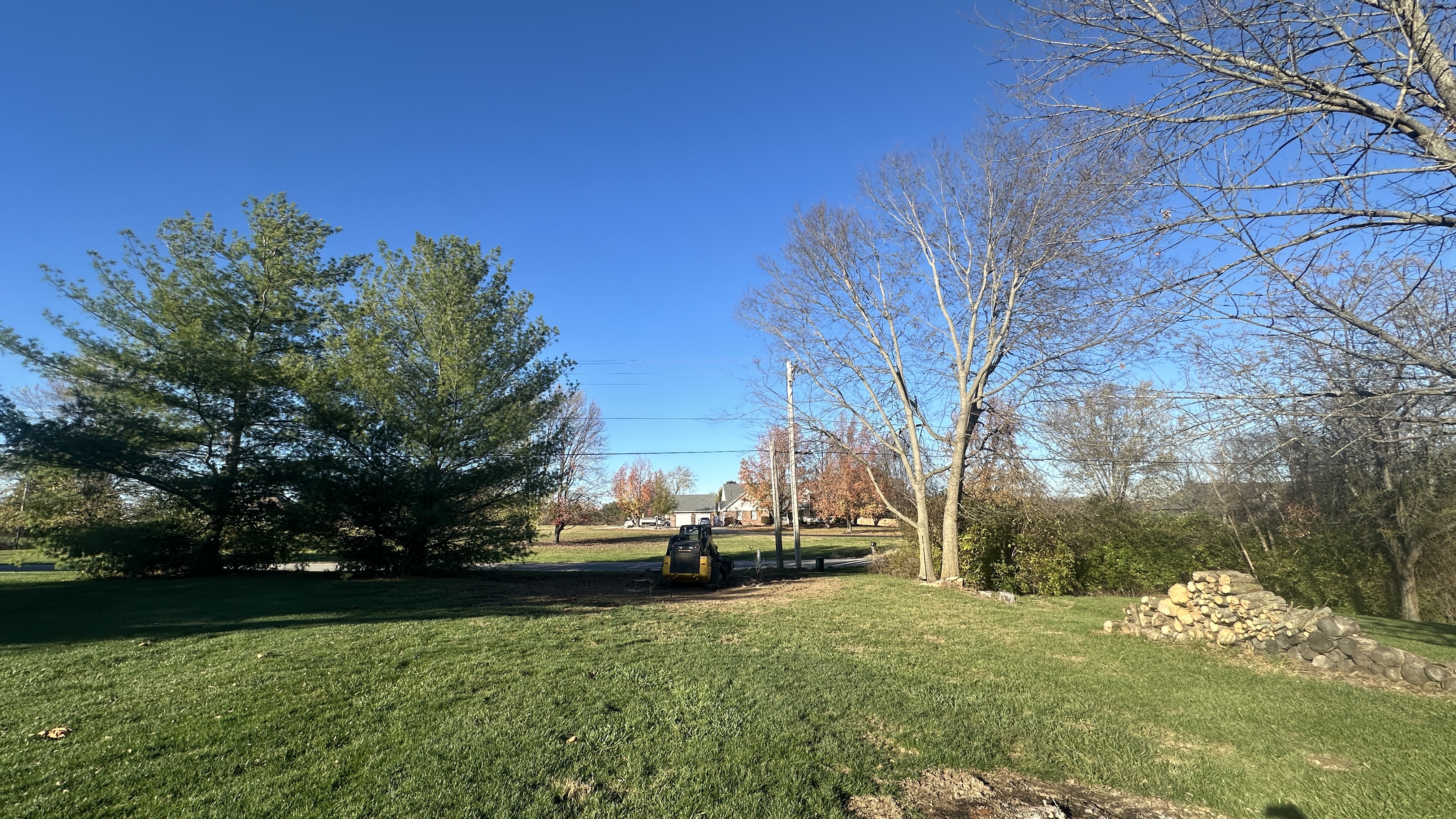 A bright, sunny day with a clear blue sky over a suburban landscape. In the foreground, a neatly manicured green lawn is partially shaded. A stack of chopped firewood is visible on the right side of the image, against a grassy slope. In the midground, a yellow compact excavator is parked on a dirt patch. Beyond that, a paved driveway leads to houses with attached garages. Several trees are scattered throughout the scene, some with green pine needles and others with bare branches, hinting at the autumn or early winter season. Power lines are visible crisscrossing the sky.