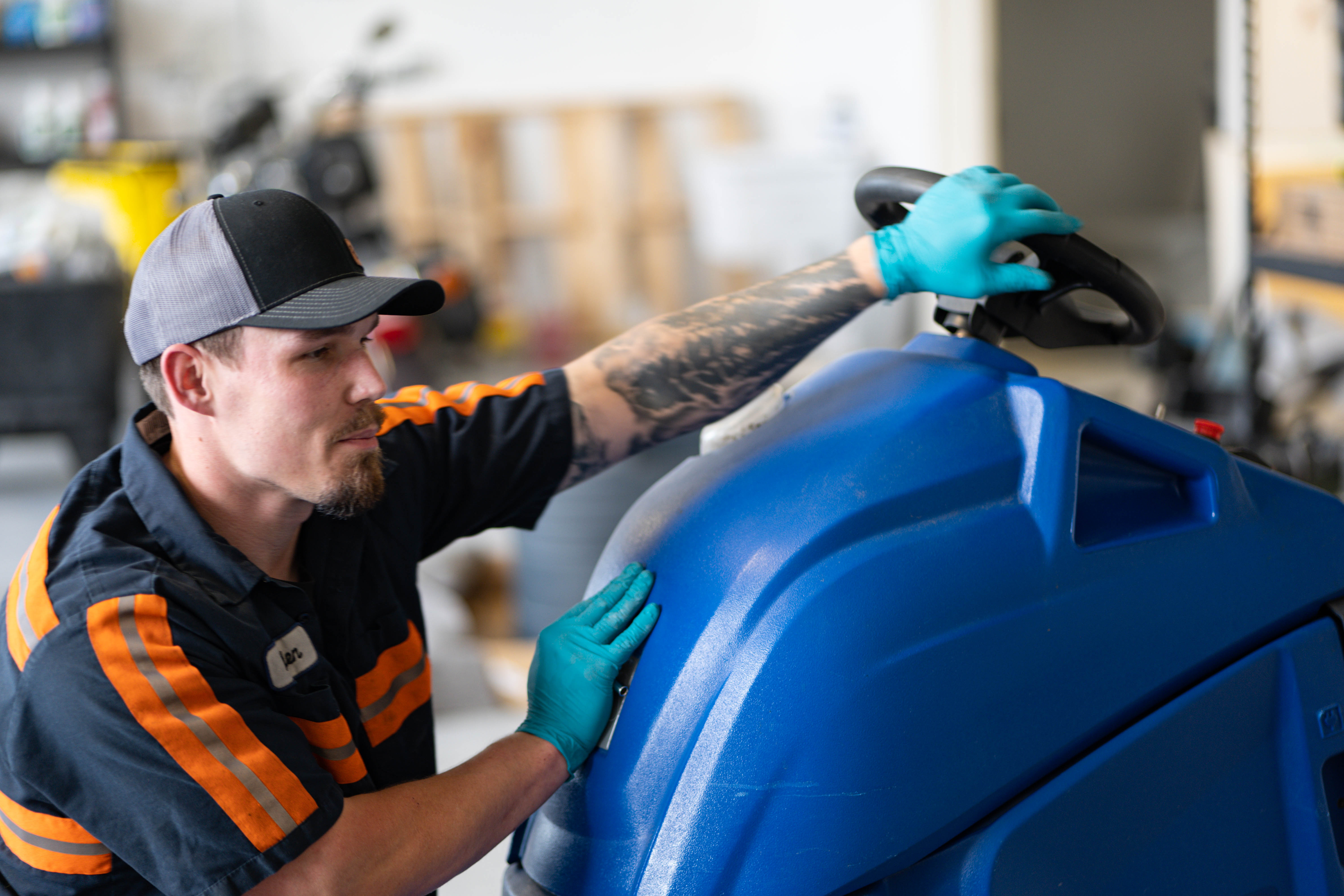 A WaySolutions service technician in a high-visibility uniform inspects the steering column and upper housing of a blue commercial floor scrubber. By providing expert on-site equipment maintenance for facilities in Layton, Utah, our team ensures precise operator control and safe handling for heavy-duty cleaning fleets