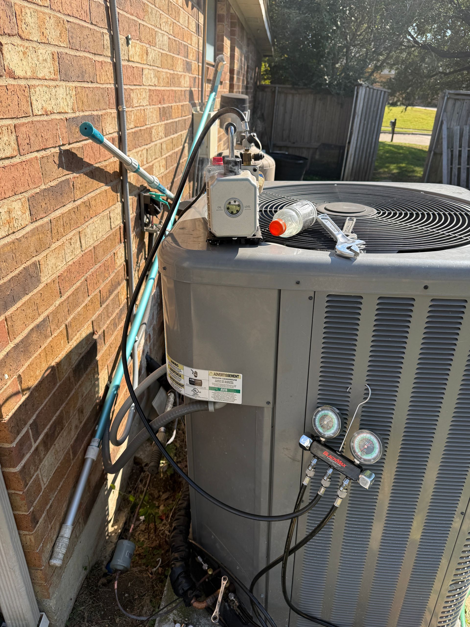Outdoor HVAC unit connected to gauges and vacuum pump during servicing. Tools and equipment placed on top, indicating refrigerant charging, diagnostics, or repair work on residential cooling system.