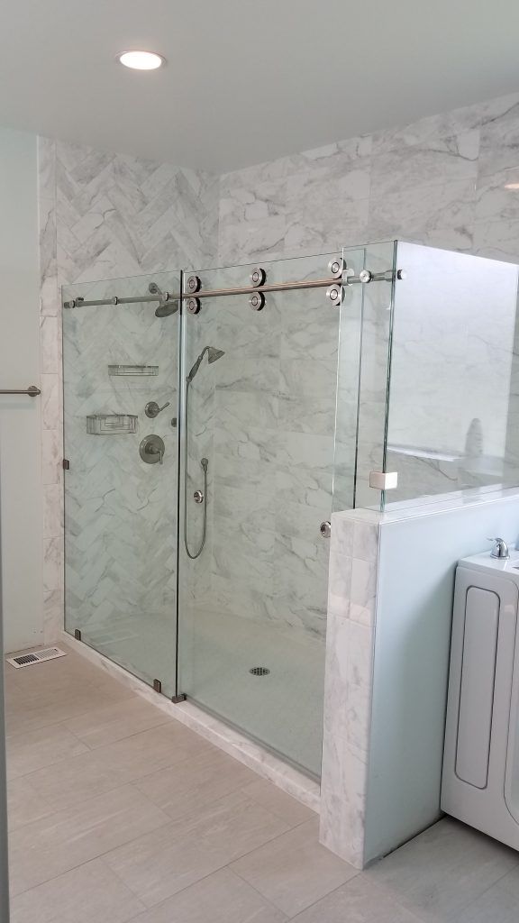 A modern bathroom featuring a large, glass-enclosed shower with chrome fixtures and marble-patterned tile walls. The shower has sliding glass doors and a rainfall showerhead. The floor is tiled with light-colored rectangular tiles. To the right of the shower is a white bathtub or washing machine.