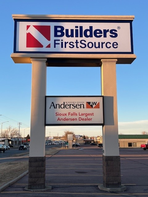 A tall street sign for "Builders FirstSource" is displayed, featuring the company's logo in red and blue. Below it is a secondary sign for "Andersen Windows & Doors," labeled as "Sioux Falls Largest Andersen Dealer." The sign is supported by two beige columns with stone bases, and the background shows a commercial street with other buildings and parked cars. The scene is lit by soft evening sunlight.