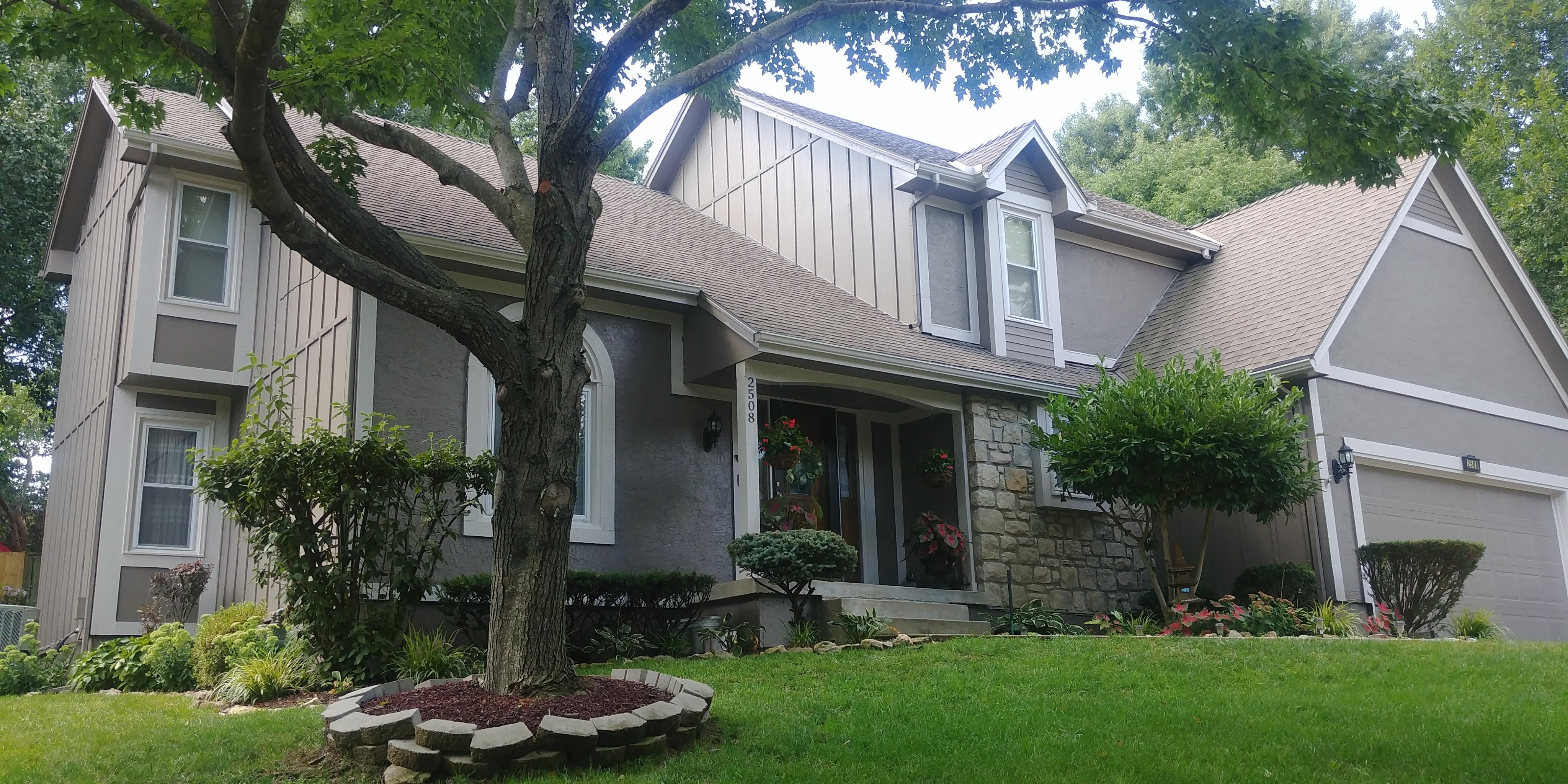 A beautifully painted two-story home featuring a fresh exterior paint job in neutral tones with white trim accents. The property showcases stone detailing, landscaped gardens, and a covered front porch, adding charm and curb appeal. Ideal for highlighting exterior painting services, trim work, and home renovation projects.