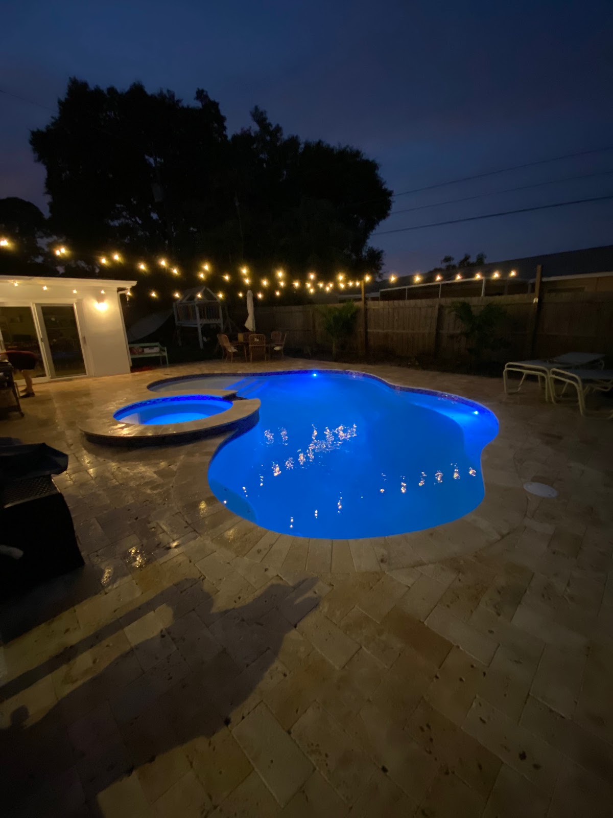 Backyard swimming pool and spa at night with bright blue underwater lighting, string lights, and stone paver decking.