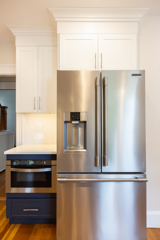 A modern kitchen features a stainless steel Frigidaire Professional French door refrigerator with an ice and water dispenser, positioned next to a built-in microwave and dark navy cabinetry. White upper cabinets with brushed nickel handles are mounted above the refrigerator and a tiled backsplash is visible behind the countertop. The kitchen flooring is made of warm-toned wood planks.