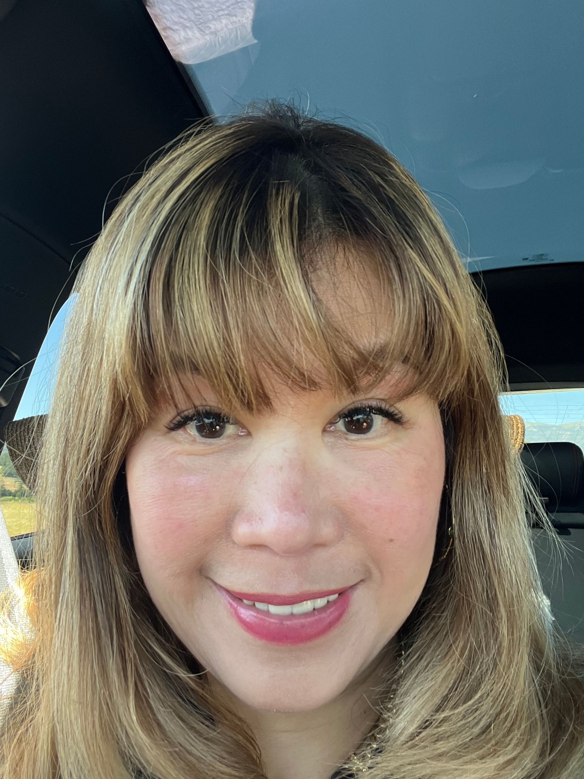 A woman smiles inside a car, with sunlight highlighting her light brown hair and bangs. She has clear skin, natural makeup, and appears relaxed while sitting in the passenger seat.