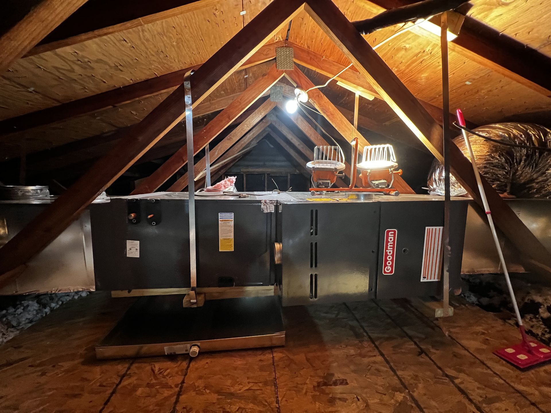 Large attic HVAC system with duct connections and lighting, showing installation or repair work. Equipment positioned across beams with visible insulation and tools, highlighting residential heating and cooling infrastructure.