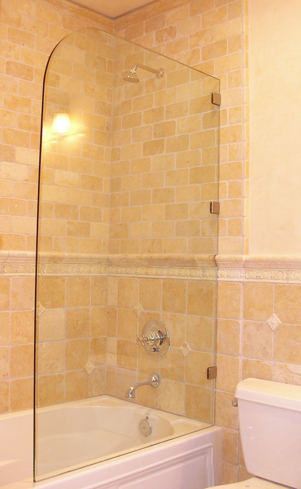 A bathroom interior featuring a bathtub with a clear glass shower screen, a showerhead, a faucet, and a toilet. The walls are tiled with small, light beige square tiles and larger, rectangular beige tiles. A decorative border runs horizontally across the wall.