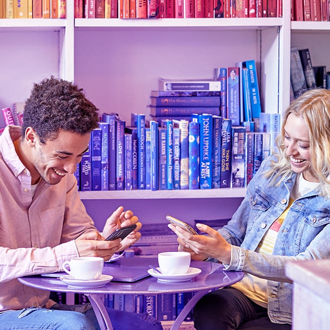 Deux personnes assises à une table violette avec des tasses de café utilisant le réseau mobile Three sur leur portable.