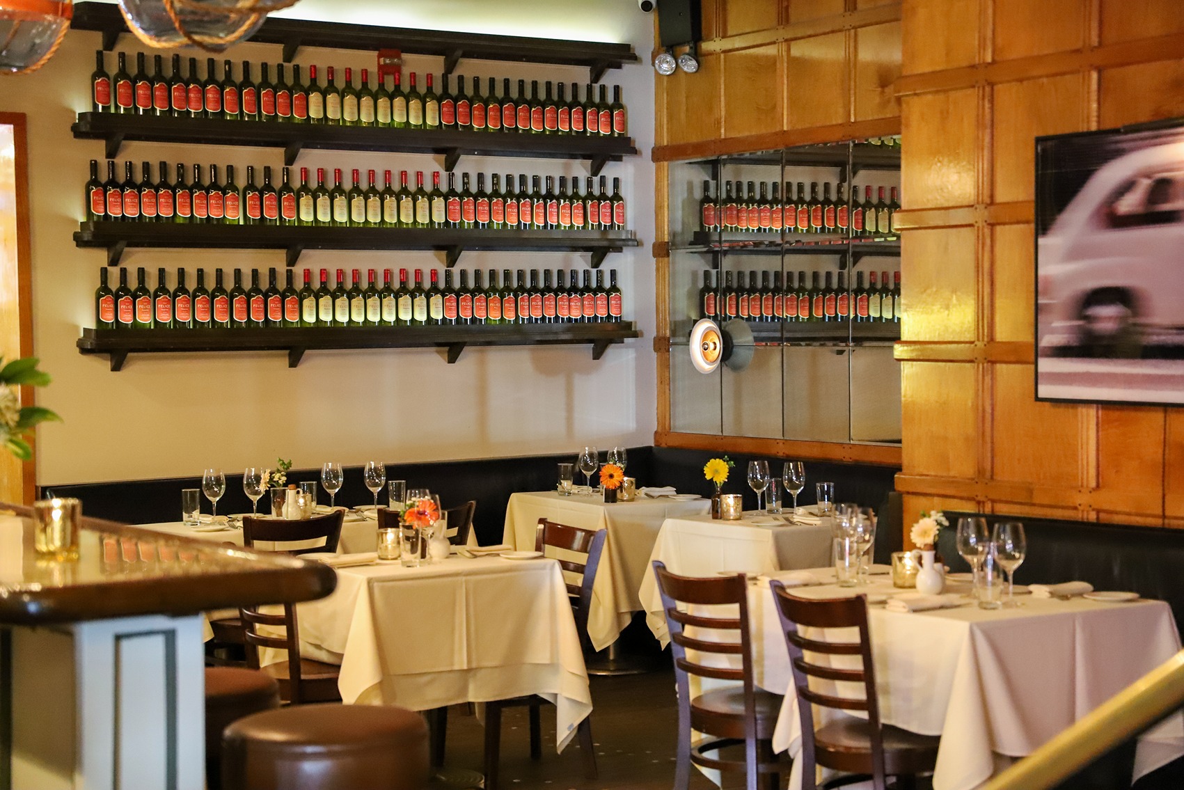Three long shelves filled with identical red-labeled wine bottles line a white wall above white-clothed dining tables. Warm wood paneling and a large mirror create a classic, inviting atmosphere.