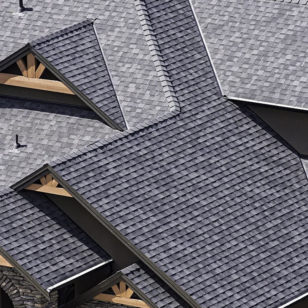 Overhead perspective highlighting the intricate angles, valleys, and gables of a modern gray shingle roof, accented with decorative wooden gable trims and stone masonry details below.