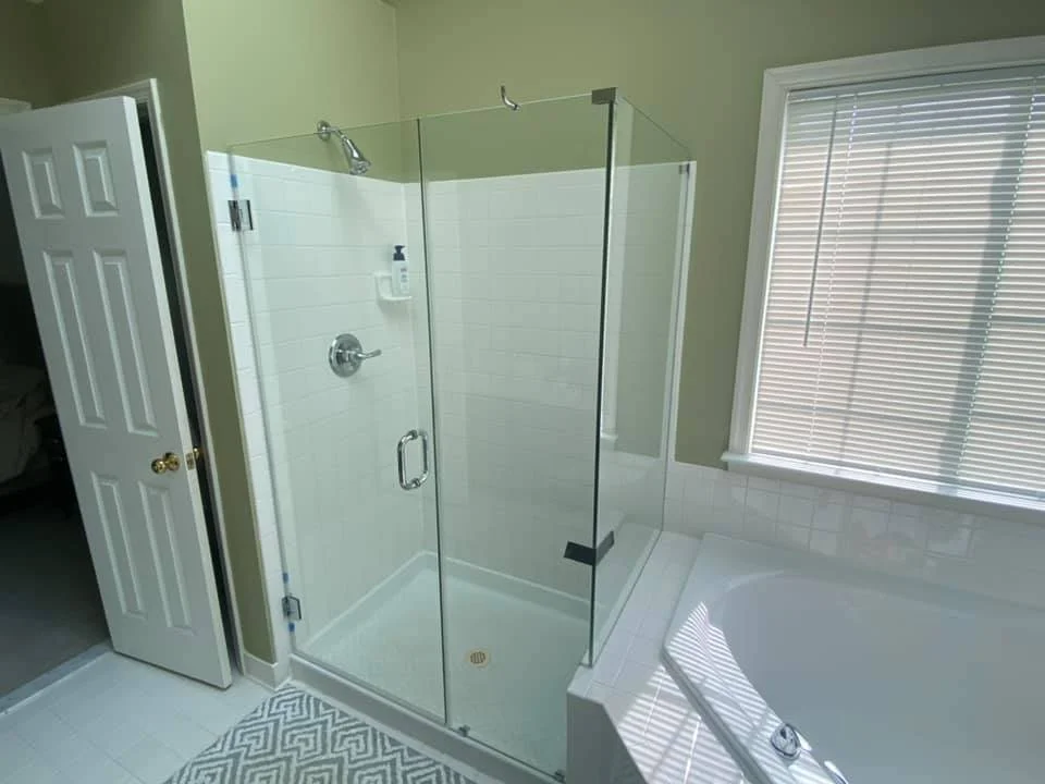 A modern bathroom featuring a spacious glass-enclosed shower and a white corner bathtub. The shower has white tiled walls and a clear glass door with a chrome handle. A shower head and a dispenser for toiletries are visible inside. The bathtub is adjacent to the shower, with a tiled surround. A window with horizontal blinds is located above the bathtub. A white door with gold hardware is open to the left of the shower, revealing a glimpse of another room. A patterned rug is on the floor in front of the shower. The walls are painted a light green color.