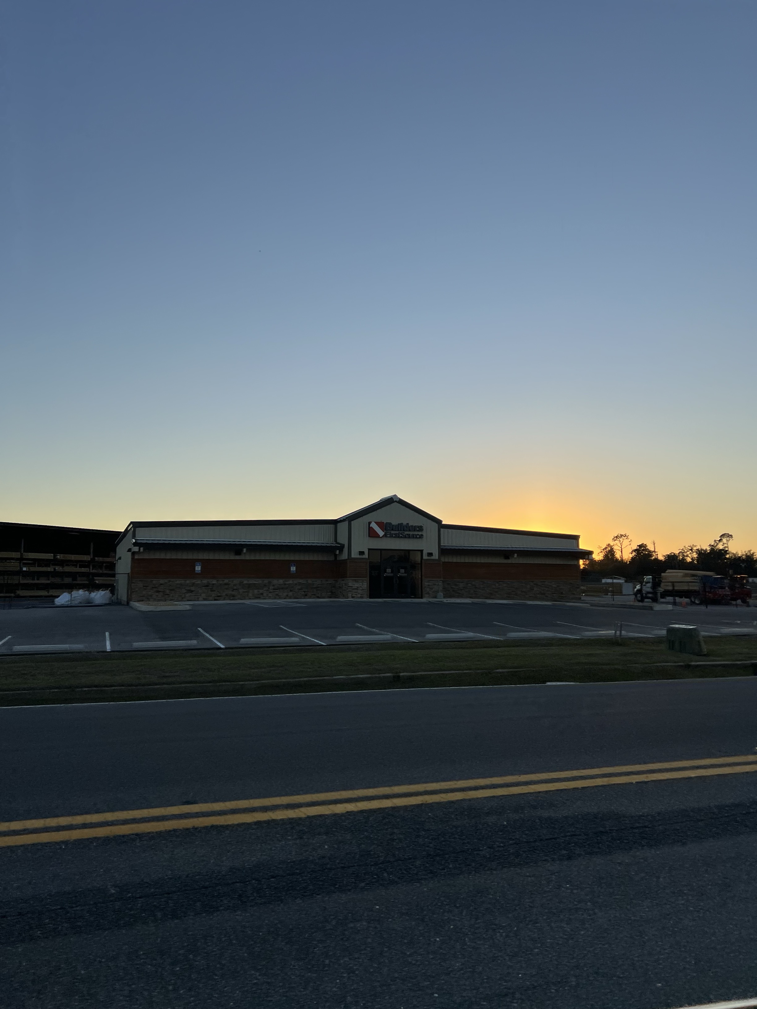 A large "Builders FirstSource" building, captured from across a road at sunset. The building has a stone and beige exterior with the company’s logo prominently displayed in the center. To the left are stacks of lumber under a covered area. The sky is clear, transitioning from light blue to a warm glow near the horizon, creating a silhouette effect on the building as the sun sets behind it.